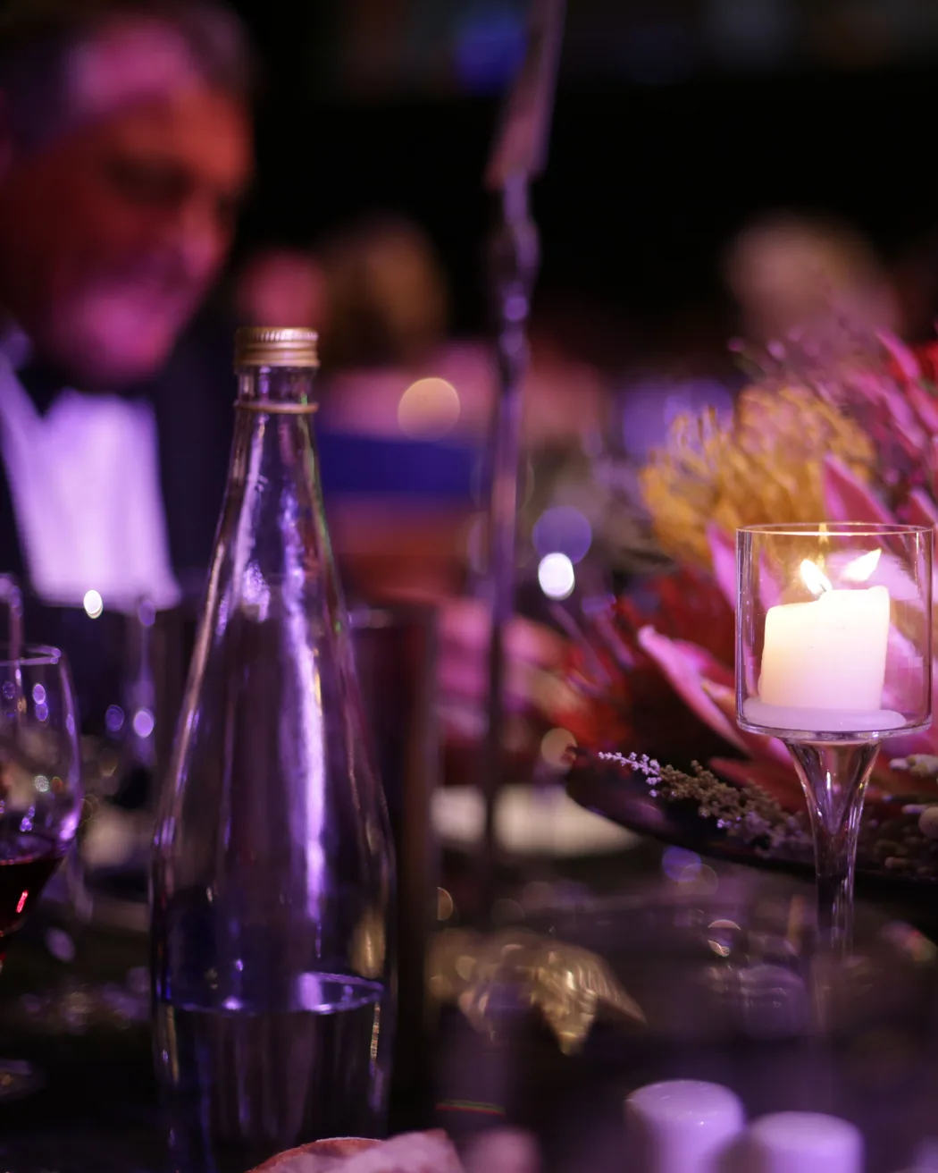 Table dressée pour un évènement privatisé, bougie et centre de table floral, bouteilles de vin et convives en arrière-plan